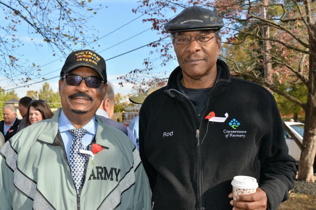 One man wearing army jacket and Vietnam Veteran hat stands beside another man in black with coffee cup in hand
